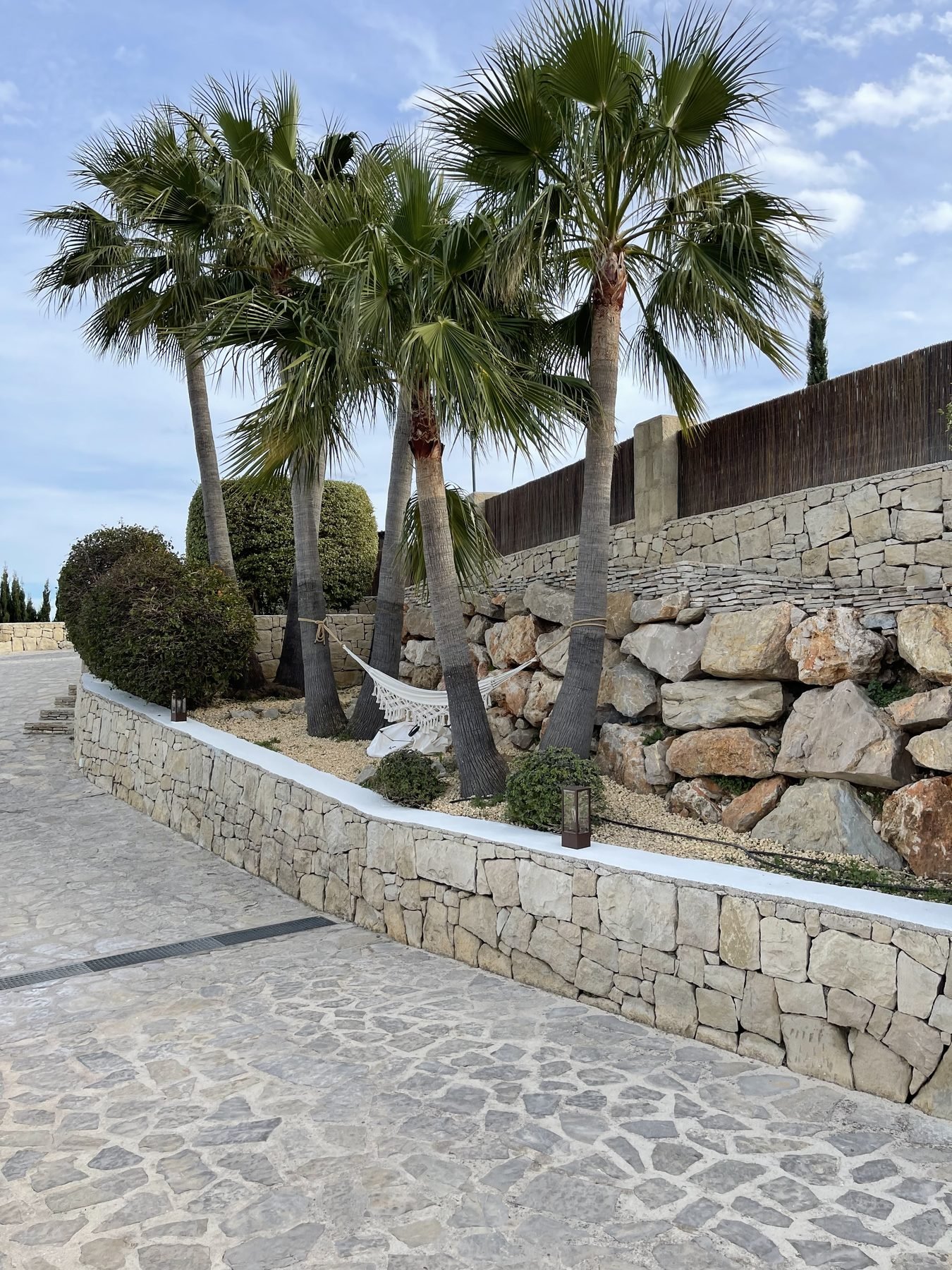 Muro de piedra natural con remate blanco entre palmeras en jardín de villa en La Sella